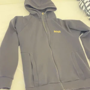 Svart hoodie med dragkedja från BOSS - Svart hoodie från BOSS med gul logga på bröstet. Tröjan har huva med snörning, hel dragkedja och två stora fickor framtill. Perfekt för chill dagar och streetstyle. Mjuk och skön känsla tack vare bomullsmaterialet.