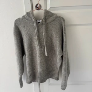 Grå stickad hoodie från Zara - Mysig grå hoodie från Zara i stickat material. Tröjan har huva med snörning, långa ärmar och breda ribbade muddar vid ärmslut och nederkant. Perfekt för dig som gillar en chill och enkel stil.