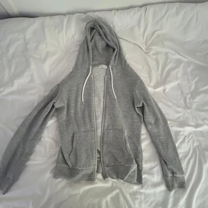 Grå hoodie med dragkedja och huva - En basic grå hoodie med dragkedja framtill, huva med vita snören och två stora fickor. Tröjan är långärmad och har ribbade muddar vid ärmslut och nederkant. Perfekt för chill dagar eller när du vill ha något bekvämt och enkelt.
