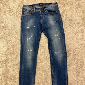 Tramarossa blå slitna jeans (slim fit) - Snygga blå jeans från Tramarossa med slitna detaljer och broderad text på insidan. Jeansen har klassisk femficksdesign, orange sömmar och knappgylf. De är tillverkade i bomull och har en smal passform som sitter riktigt bra.