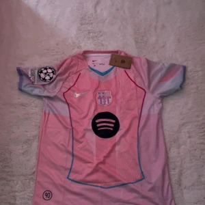 Barça x Nike rosa fotbollströja - Säljer en FC Barcelona x Nike fotbollströja i rosa och ljusblått med korta ärmar. Tröjan har klubbmärke, Nike-logga, Spotify-sponsor och Champions League-märke på ärmen. Tillverkad i Dri-FIT-material som andas. Snygga kontrastdetaljer i blått och rött.