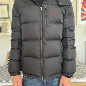 Svart pufferjacka Polo Ralph Lauren XL - Svart pufferjacka från Polo Ralph Lauren i storlek XL jag är 174cm. Jackan har en klassisk quiltad look, dragkedja framtill och två sidofickor med dragkedja. Ikoniska röda Polo-loggan på bröstet. Perfekt för kalla dagar och riktigt snygg till jeans.