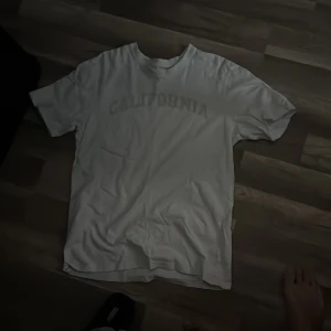 Vit t-shirt med California-tryck - Vit t-shirt med trycket 'CALIFORNIA' i ljus ton över bröstet. Klassisk rund hals och korta ärmar, perfekt för chill dagar. Materialet känns som mjuk bomull och passformen är normal. Enkel och clean stil som funkar till jeans eller shorts.