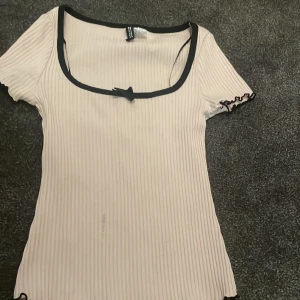 Ribbad ljusrosa topp med rosett H&M - Supersöt ljusrosa ribbad topp från H&M Divided med svart kant runt den fyrkantiga urringningen och en liten rosett framtill. Korta ärmar med vågig svart kant och figurnära passform. Perfekt för en trendig och söt look.