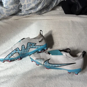 Nike Air Zoom fotbollsskor vit/blå - Snygga Nike Air Zoom fotbollsskor i vitt med blå och svarta detaljer. Skorna har låg profil, mönstrad sula för bra grepp och en stilren design med stora Nike-loggor på sidorna. Perfekta för dig som vill sticka ut på planen.