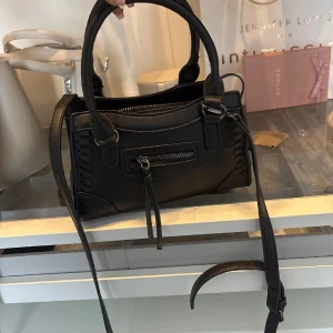 Svart handväska med dragkedja - Snygg svart handväska i skinnimitation med både handtag och lång axelrem. Väskan har dekorativa sömmar på sidorna och en praktisk dragkedjeficka framtill med hängande detaljer. Perfekt storlek för vardagens essentials.