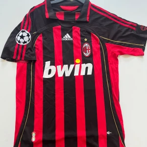 AC Milan Kaka #22 Adidas fotbollströja - Säljer en klassisk AC Milan fotbollströja från Adidas med Kaka och nummer 22 på ryggen. Tröjan är röd och svart med vertikala ränder, Champions League-märke på ärmen och guldfärgade detaljer. Materialet är lätt och andas, perfekt för match eller träning.