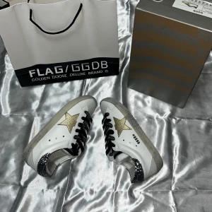 Golden Goose sneakers med glitter - Golden Goose sneakers i vitt skinn med guldstjärna på sidan och svart snörning med logotyp. Hälpartiet har glittriga silverdetaljer som ger extra edge. Snygg platt sula och rund tå, perfekt för dig som gillar att sticka ut med coola detaljer.