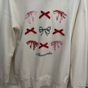 Vit hoodie med rosettprint Abercrombie - Vit hoodie från Abercrombie Kids med tryck av röda och rosa rosetter samt en grå rosett i mitten. Tröjan har huva och lång ärm, samt ribbade muddar vid ärmslut och nederkant. Perfekt för dig som gillar söta detaljer och comfy stil.