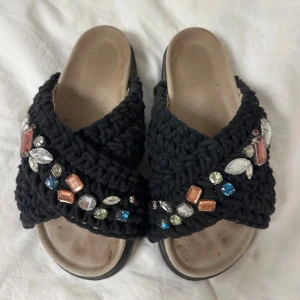 Svarta sandaler med pärlor från Inuikii - Unika svarta sandaler från Inuikii med grovstickade band och färgglada pärlor och stenar i olika former. Sandalerna har platt sula och öppen tå, vilket gör dem riktigt chill och snygga. Perfekt för dig som gillar statement-detaljer och vill sticka ut.