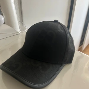 Svart keps från Gucci med GG-mönster - Snygg svart keps från Gucci med diskret GG-mönster över hela kepsen. Klassisk böjd skärm med läderdetalj längs kanten och justerbar rem bak. Tillverkad i bomull och polyester för en lyxig känsla och passform.