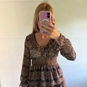 Brun mönstrad kortklänning med volang - Supersöt kortklänning i brunt och svart med blommigt mönster. Klänningen har långärmade transparenta ärmar, smockad midja och flera volanger i kjolen. V-ringad hals och lätt, luftig känsla. Perfekt för hösten! 