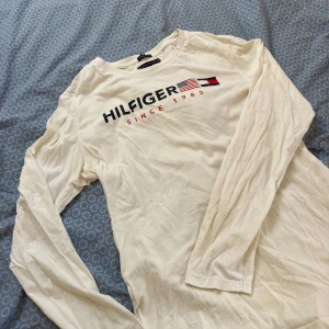 Vit långärmad tröja Tommy Hilfiger - Vit långärmad tröja från Tommy Hilfiger med tryck framtill och flaggdetaljer. Tillverkad i ekologisk bomull, rund halsringning och klassisk passform. Säljer då den inte kommer till användning.