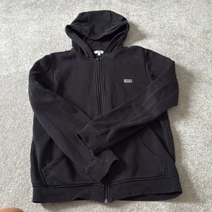 Svart hoodie från BOSS med dragkedja - Svart hoodie från BOSS med diskret logga på bröstet. Tröjan har huva, långa ärmar och praktiska fickor framtill. Tillverkad i mjuk bomull och stängs med dragkedja. Perfekt för chill dagar eller när du vill ha en clean streetstyle-look.