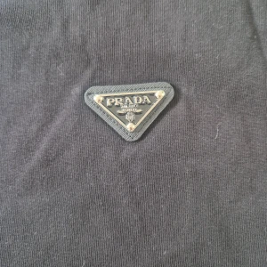 Prada tshirt - Svart t-shirt från Prada med klassisk rund hals och korta ärmar. På bröstet sitter en ikonisk triangulär Prada-logga i metall. T-shirten är gjord i mjuk bomull och har en clean, stilren look som passar perfekt till jeans eller shorts.