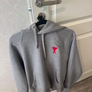 Grå hoodie från Ami Paris - Säljer en grå hoodie från Ami med klassisk huva och snörning. På bröstet finns Ami:s ikoniska röda hjärta med A, och en diskret Ami-brodering på fickan. Tröjan är i mjuk bomull och har en normal passform med långa ärmar. Perfekt för chill dagar. 
