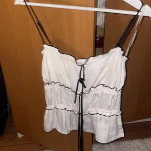 Ny gina topp - Supersöt vit linnetopp med svarta tunna axelband och svarta sömmar längs kanterna. Toppen har volangkant nertill och en snygg knytning framtill. Perfekt för varma dagar och ger en trendig vibe.