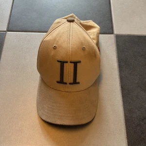 Beige keps från Les Deux - Snygg beige keps från Les Deux med broderad II-logga framtill i mörkgrönt. På sidan finns små broderade ränder i blått, vitt och rött. Justerbar snapback baktill och tillverkad i en mix av bomull och polyester. Perfekt accessoar för en clean och stilren look.