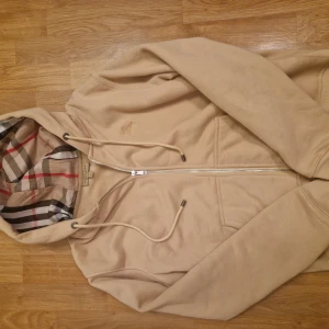 Beige hoodie från Burberry med dragkedja - Snygg beige hoodie från Burberry med klassiskt rutigt foder i huvan och broderad logga på bröstet. Tröjan har hel dragkedja, snörning i huvan och är tillverkad i mjuk bomull. Perfekt för dig som gillar stilrena och exklusiva plagg. Pris kan diskuteras