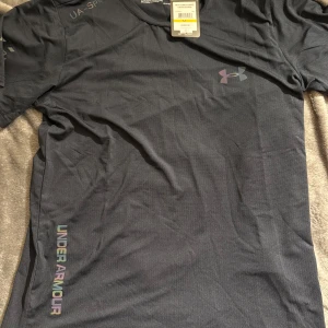 Svart Under Armour HeatGear t-shirt - Svart t-shirt från Under Armour med HeatGear-teknologi som håller dig sval och torr. Klassisk rund hals, korta ärmar och reflekterande logga på bröstet. Tillverkad i lätt och snabbtorkande polyester, perfekt för träning och löpning. Snyggt tryck med Under Armour längs sidan. STOLREK M men det Passar perfekt för S 