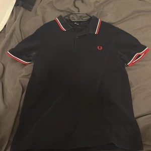 Svart Fred Perry t-shirt med röd/vit kant - Svart t-shirt från Fred Perry i storlek L med klassisk polokrage och korta ärmar. Snygga röda och vita ränder på krage och ärmslut, samt Fred Perry-logga broderad i rött på bröstet. Tillverkad i mjuk bomull som känns skön mot huden.