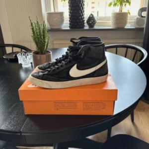 Nike Blazer Mid svart/vit sneakers - Säljer ett par klassiska Nike Blazer Mid sneakers i svart mocka med vit swoosh och vit sula. Skorna har snörning och en retro vibe med högt skaft. Perfekt för dig som gillar streetstyle och vill ha en ikonisk siluett i garderoben.