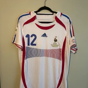 Frankrike Henry VM 2006 tröja Adidas - Säljer en Frankrike fotbollströja från VM 2006 med Henry och nummer 12 på ryggen. Tröjan är vit med blå och röda detaljer, Adidas-logga på bröstet och FIFA World Cup-patch på ärmen. Tillverkad i lätt polyester, kortärmad och med snygga grafiska linjer framtill.