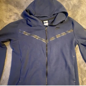 Marinblå Nike hoodie med dragkedja - Snygg marinblå hoodie från Nike med hel dragkedja och huva. Tröjan har svarta detaljer över bröstet och en praktisk ficka med dragkedja på ärmen. Perfekt för dig som gillar sportig och avslappnad stil.