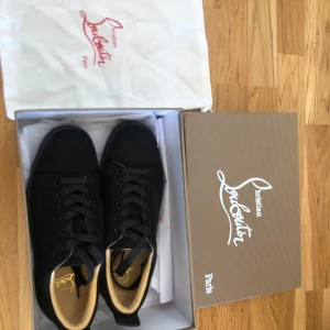 Svarta sneakers från Christian Louboutin - Stilrena svarta sneakers från Christian Louboutin med klassisk röd sula och broderad logga på hälen. Skorna har snörning, rund tå och är tillverkade i mocka med beige innersula. Perfekta för dig som vill ha en lyxig och clean look. Inga prutningar tar jag emot tyvärr!