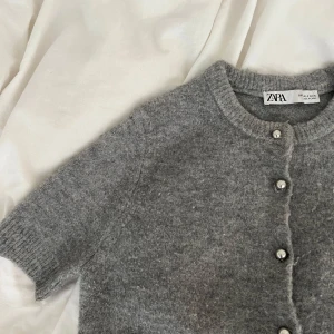 Grå stickad kofta från Zara - Mysig grå stickad kofta från Zara med korta ärmar och rund hals. Koftan har silvriga knappar framtill och ribbade muddar vid ärmslut och nederkant. Perfekt för lager-på-lager och enkel att styla till olika looks.