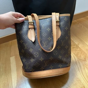 Louis Vuitton Monogram axelväska - Säljer en klassisk Louis Vuitton axelväska i brunt monogrammönster med ljusbruna skinnhandtag och detaljer. Väskan har justerbara axelremmar, guldfärgade metalldetaljer och dragkedja på insidan. Perfekt storlek för vardagens essentials. Autentitetsbevis finns från Vestiaire Collective.