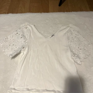 Vit blus med spetsärmar och v-ringning - Supersöt vit blus med v-ringad hals och korta ärmar i blommig spets. Ärmarna är vida och har ett detaljerat, utstansat blommönster som ger en romantisk vibe. Perfekt att styla med jeans eller kjol för en fräsch look.