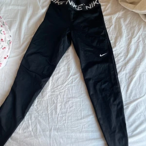Svarta Nike leggings med logga - Säljer ett par svarta Nike leggings med bred resår i midjan där det står NIKE i vitt. Snygg, tight passform och klassisk Nike-logga på benet. Perfekta för träning eller chill, tillverkade i stretchigt syntetmaterial som sitter skönt och följer kroppen.