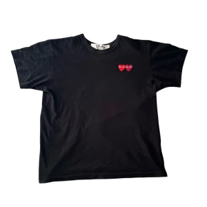 Svart t-shirt från Comme des Garçons - Säljer en svart t-shirt från Comme des Garçons med två röda hjärtan med ögon broderade på bröstet. T-shirten har en normal passform och är kortärmad. Perfekt för en stilren look! Det står storlek M men skulle säga att den sitter mer som S