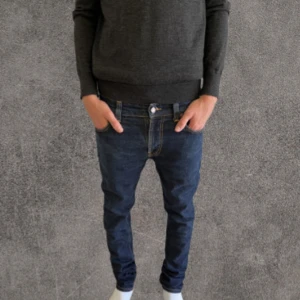 Mörkblå slim jeans från Nudie Jeans - Säljer ett par mörkblå jeans från Nudie Jeans med snygga kontrastsömmar och klassisk broderad bakficka. Modellen har normal passform och raka ben, tillverkade i slitstarkt bomullsmaterial. Perfekt för dig som gillar stilrena och tidlösa jeans.