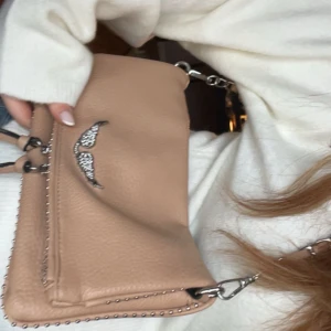 Beige axelväska med silverdetaljer - Snygg beige axelväska i skinn med silverkedja och dekorativa vingar i strass på framsidan. Väskan har dubbla dragkedjor och små nitar längs kanterna som ger en cool touch. Perfekt storlek för mobilen och det viktigaste.