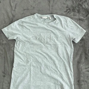 Ljusgrå GANT t-shirt med logga - Snygg ljusgrå t-shirt från GANT med broderad logga och texten 'American Sportswear' framtill. Klassisk rund hals och korta ärmar. Perfekt för en chill och stilren look, tillverkad i mjuk bomull som känns skön mot huden.