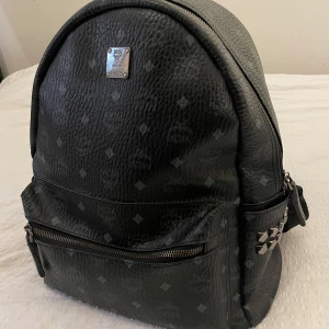 MCM BACKPACK/RYGGSÄCK - Black - Höj din stil med denna MCM-ryggsäck med ett genomgående Monogramtryck. Utrustad med en toppstängning, dekorativa guldtonade nitar och flera fickor för dina väsentligheter. Tillverkad av en blandning av bomull och polyuretan, är denna ryggsäck både stilfull och funktionell. Väldigt limiterad och exklusiv modell slutsåld överallt. Nypris 14 145kr på Miinto.se, säljes nu för enbart 7999kr.  Mått: Höjd: 37 cm   Längd: 29 cm   Vidd: 14 cm   