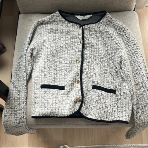 Vit och svart kofta från H&M - Snygg kofta från H&M i vit och svart med strukturerat rutigt mönster. Svarta detaljer vid kanter och fickor, samt guldfärgade knappar framtill. Långärmad och perfekt för lager-på-lager. Passar dig som gillar klassisk stil med en twist.har lite för på som inte syn mycket när man har på den.