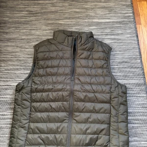 Mörkgrön quiltad dunväst - Snygg mörkgrön dunväst med quiltad design och hög krage. Västen har en rak passform och dragkedja framtill. Perfekt att slänga över en hoodie eller tröja när det är lite kyligt ute. Materialet är lätt och känns skönt mot kroppen. Storlek M.