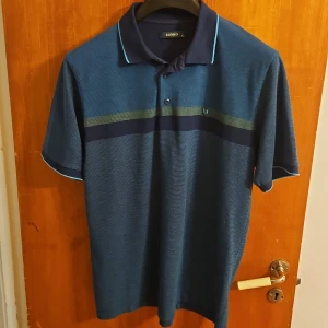 Blå pikétröja från Konko XL - Snygg blå pikétröja från Konko i storlek XL. Tröjan har korta ärmar, krage med knappar och en diskret randig detalj i grönt och mörkblått över bröstet. Perfekt för en chill och stilren look. Materialet känns som bomull och är bekvämt mot huden.