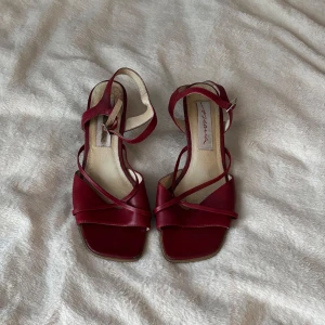 Röda sandaler med klack och remmar - Snygga röda sandaler med smala remmar och fyrkantig tå. Skorna har en låg klack i trälook och spänne runt vristen.