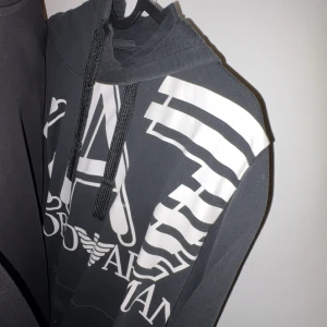 Svart hoodie från Armani med tryck - Snygg svart hoodie från Armani med stort vitt grafiskt tryck och logga framtill. Tröjan har huva med svarta snören och är tillverkad i mjukt bomullsmaterial. Perfekt för en avslappnad och trendig look.