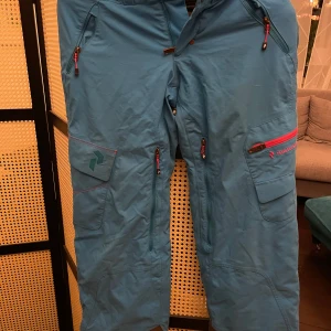 Blå täckbyxor Peak Performance - Snygga blå täckbyxor från Peak Performance med rosa dragkedjor och detaljer. Byxorna har flera fickor, lågmidjade och broderad logga. Perfekta för vinterns äventyr och outdoor-aktiviteter. Materialet är slitstarkt och känns sportigt.