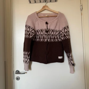 Brun och ljusrosa stickad kofta - Supermysig stickad kofta i brunt och ljusrosa med snyggt mönster över bröst och ärmar. Koftan har stora knappar framtill och ribbade muddar. Perfekt för lager-på-lager och chill höstkvällar. Från märket Odd Molly. Storlek 2 som enligt deras storleksguide motsvarar en storlek M. Fint skick dock kan koftan ha ytludd och noppror för att materialet är ull och nylon. 