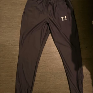 Svart zip-tröja från Under Armour och tränings byxa - Säljer en svart långärmad zip-tröja från Under Armour med vit logga på bröstet. Tröjan har hel dragkedja framtill och är gjord i ett mjukt syntetmaterial som passar perfekt till träning. Snygg och sportig look med normal passform. Säljer den dessutom med en flexibel tränings byxa.