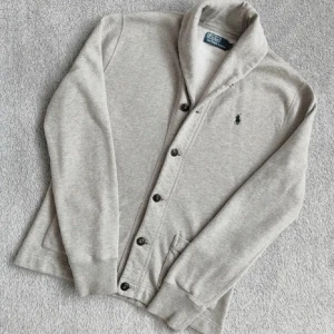 Grå kofta från Polo Ralph Lauren - Snygg ljusgrå kofta från Polo Ralph Lauren med klassisk krage och svarta knappar. Broderad logga på bröstet och två fickor framtill. Tillverkad i mjuk bomull, perfekt för lager-på-lager under höst och vår.