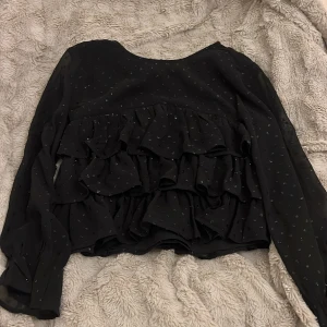 Svart volangblus från Gina Tricot - Supersöt svart blus från Gina Tricot med glittriga prickar och flera lager volanger framtill. Långärmad modell i lätt och luftig polyester, perfekt när du vill sticka ut lite extra. Blusen har rund hals och transparenta ärmar för en cool look.