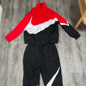 Nike - set retro  - Nike-set i färgstark röd, svart och vit färgblock-design. Jackan har dragkedja framtill, hög krage och elastiska muddar. Byxorna har 2 fickor och dragsko i midjan. Materialet är lätt och syntetiskt, perfekt för blåsiga dagar. Snygg streetstil med stor vit swoosh-detalj över bröstet. Aldrig använd, säljs pga fel storlek. Nypris 1299. 