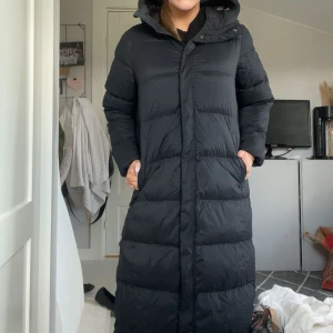 Svart lång dunjacka Mckinley - Säljer en svart lång dunjacka med huva och dragkedja. Jackan är quiltad och har en clean look med raka linjer och praktiska fickor på sidorna. Perfekt för kalla dagar när du vill hålla dig varm och ändå se stilren ut. pris går att diskutera!!